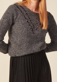 Grauer Strickpullover mit taillierter Silhouette, der mit einem V-förmigen Ausschnitt verziert ist, der mit Pailletten geschmückt ist. Kombiniert mit einem gefalteten schwarzen Rock.