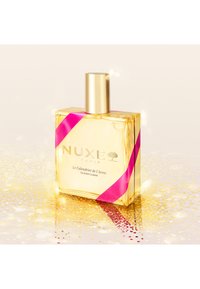 Bottiglia di profumo NUXE con un design rettangolare dorato, un tappo dorato e un accento di nastro rosa, posizionata su uno sfondo dorato scintillante.