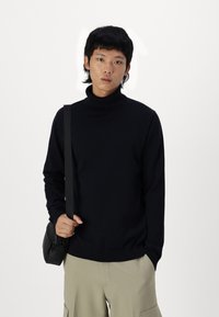 Suéter de cuello alto navy en un material de punto suave, con un diseño ajustado y puños acanalados, combinado con pantalones cargo de color beige claro y un bolso negro.