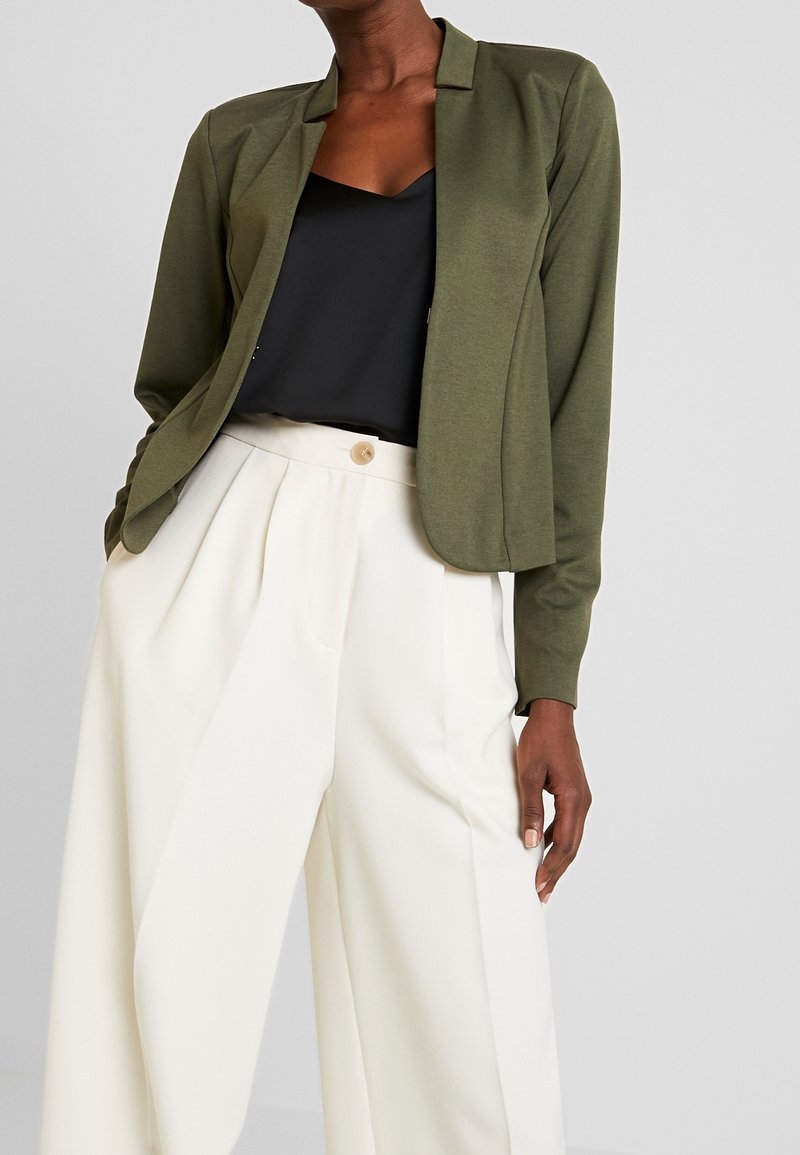 Personne portant un blazer vert olive, un haut noir et un pantalon blanc large taille haute avec un bouton à la taille, sur un fond uni.
