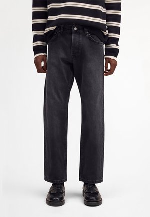 Osoba ubrana w czarne, proste dżinsy, czarne mokasyny z frędzlami oraz czarno-biały sweter w paski z długim rękawem.