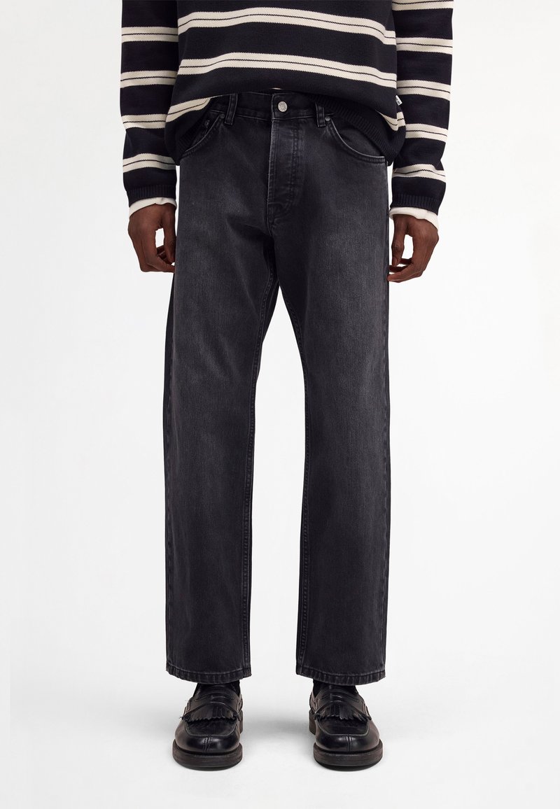 Henkilö, jolla on mustat suorahousuiset farkut, mustat hapsulliset loafersit ja mustavalkoraidallinen pitkähihainen villapaita.