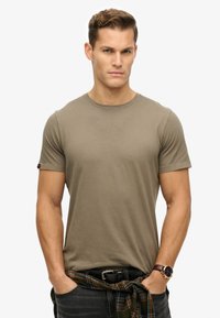 Olijfgroene katoenen t-shirt met een ronde hals en korte mouwen, gedragen met donkere jeans en een geruite shirt om de taille geknoopt.