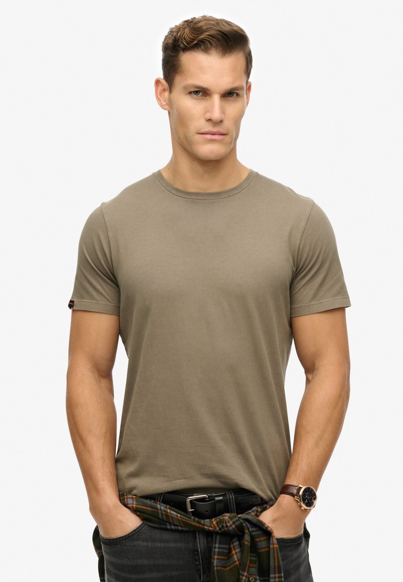 Olijfgroene katoenen t-shirt met een ronde hals en korte mouwen, gedragen met donkere jeans en een geruite shirt om de taille geknoopt.