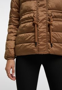 Giacca imbottita marrone con finitura lucida, dotata di cordini in vita, tessuto testurizzato e tasche laterali. Indossata sopra leggings neri.