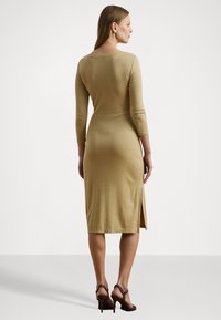 Robe en tricot légère couleur beige, longueur genou, à manches longues. Elle présente une fente dans le dos discrète et une silhouette ajustée, accompagnée de sandales à talons hauts.