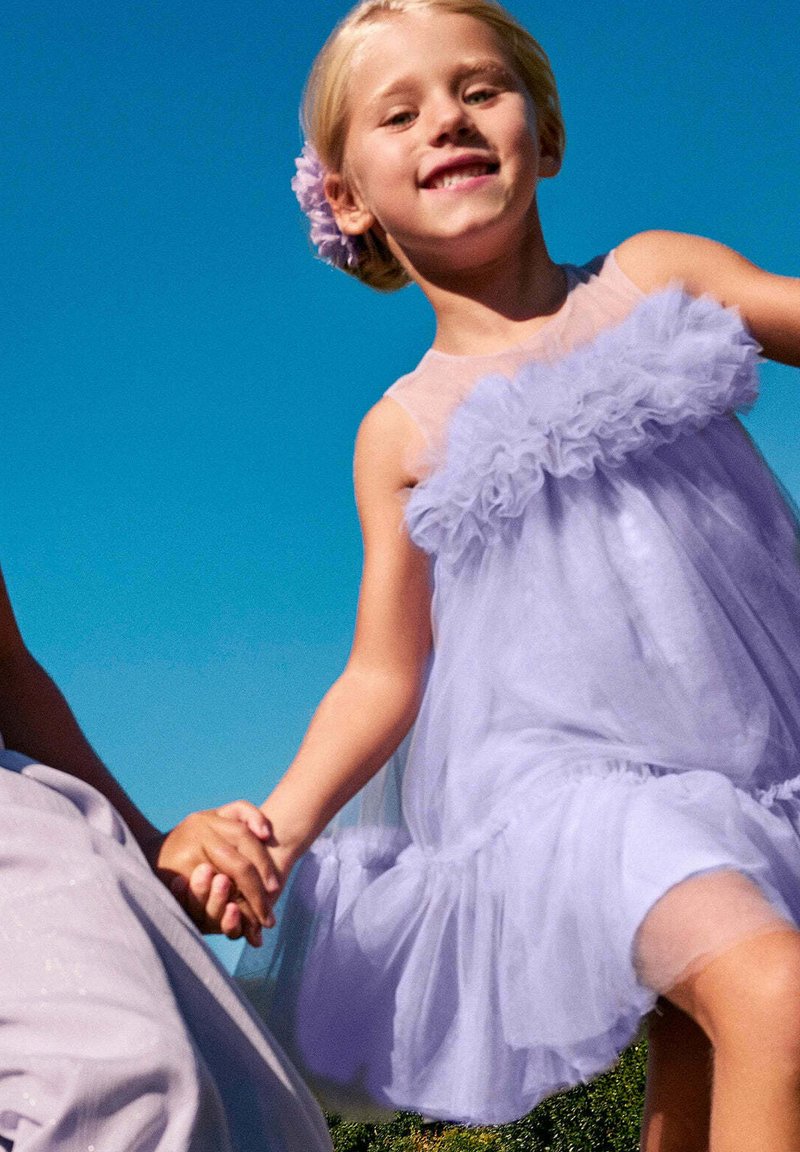 Abito di tulle lilla con un corpetto trasparente e un colletto arricciato, abbinato a un accessorio floreale nei capelli, mentre teneva per mano un altro bambino.