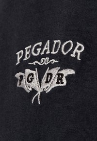 Zwarte stof met een gestructureerde afwerking, met witte geborduurde tekst "PEGADOR" en twee gestileerde vlaggen met de initialen "PG" en "DR".