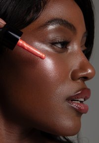 Flüssiger Highlighter wird mit einem korallenfarbenen Applikator aufgetragen. Die Haut hat ein glattes, strahlendes Finish in warmen Tönen, das die Highlights der Wangenknochen betont.