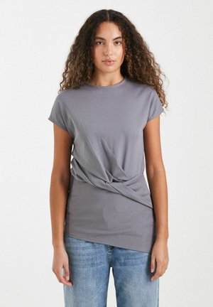 Mujer con cabello largo y rizado, vestida con una camiseta gris de manga corta con un detalle de giro delantero y vaqueros azul claro, posando delante de un fondo liso.