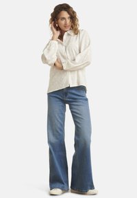 Chemise blanche à manches longues avec un motif gaufré subtil, associée à un jean flare bleu clair et des baskets blanches. Le mannequin pose avec la main dans les cheveux.