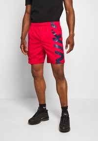 Shorts de sport rouges avec une ceinture élastique, arborant un grand logo Nike bleu marine sur le côté. Fabriqués en tissu léger et lisse.