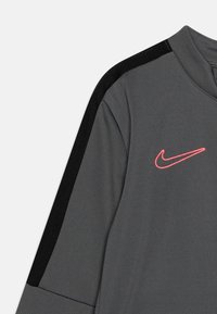Grå atletisk skjorte med høj krave, sort accent på skulderen og et lyserødt broderet Nike-logo. Stoffet er glat med en let tekstur.
