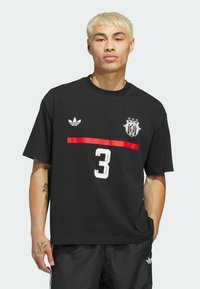 Black cotton T-shirt with short sleeves, featuring a large white number "3" and a red horizontal stripe. Adidas logo on the left chest.