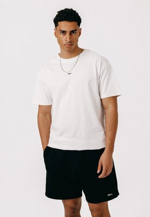 Ung mand med kort krøllet hår iført en simpel hvid t-shirt, sorte shorts og en sølvkæde, står mod en hvid baggrund.