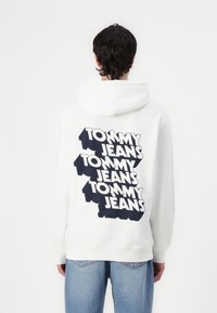 Hoodie blanc avec un grand texte "TOMMY JEANS" en noir au dos, de coupe normale et avec une poche kangourou à l'avant.