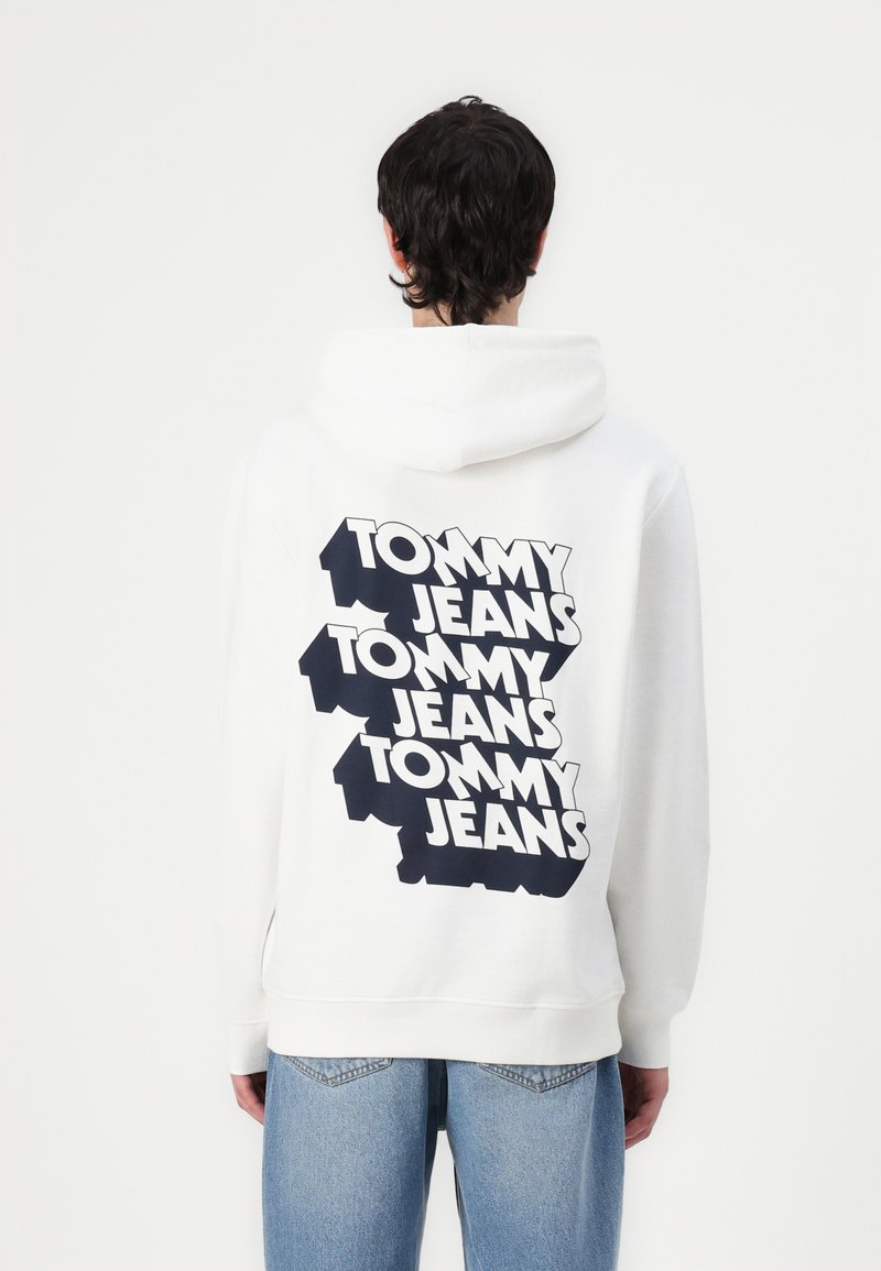 Hoodie blanc avec un grand texte "TOMMY JEANS" en noir au dos, de coupe normale et avec une poche kangourou à l'avant.