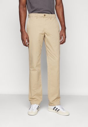 Homme portant un pantalon beige droit, un t-shirt gris et des baskets blanches à rayures noires, debout devant un fond uni.