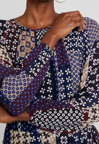 Personne à la peau foncée portant un chemisier patchwork à manches longues avec des motifs géométriques bleus, noirs, blancs et beiges, les bras croisés.