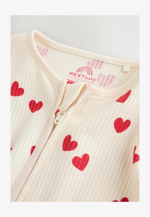 Tutina per neonati color crema a coste con motivi a cuoricini rossi, dotata di zip frontale e scollatura morbida. L'etichetta riporta la scritta "NEXTbaby" con un logo arcobaleno.