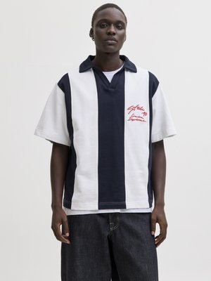 Mladić odjeven u polo majicu s kratkim rukavima, tamnoplavu i bijelu s okomitim prugama, s crvenim tekstom na prsima, u kombinaciji s tamnim širokim trapericama.