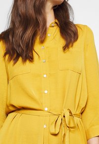 Abito giallo con bottoni, dotato di una cintura in vita, due tasche sul petto, maniche corte e un tessuto texturizzato. Chiusura semplice con bottoni.