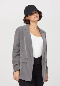 Blazer grigio con revers, un taschino frontale e abbinato a una maglietta bianca e leggings neri. Un berretto nero completa il look.