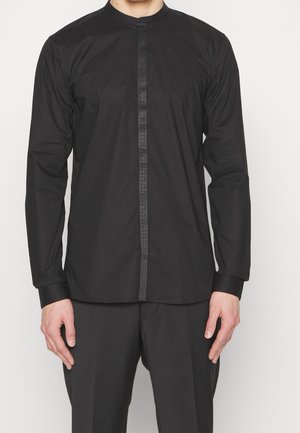 Homme portant une chemise noire à manches longues avec une bande décorative verticale le long de la ligne de boutons, associée à un pantalon noir.