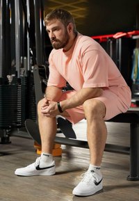 Outfit sportivo di colore rosa chiaro con maniche corte e pantaloni corti; design semplice. Sneakers bianche con accenti neri; ambiente di palestra con attrezzature sullo sfondo.
