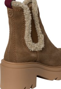 Braune Wildleder-Stiefelette mit einem flauschigen beigen Kunstfellkragen, elastischem seitlichem Einsatz und einer klobigen hellbraunen Sohle mit strukturiertem Grip.