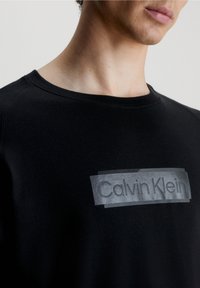 Svart sweatshirt med rund hals, med en blank grå Calvin Klein-logotyp på en genomskinlig lapp. Slät textur, modern passform.