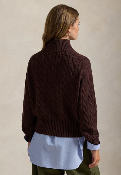 Hnedý sveter s vrkočovým vzorom, vysokým golierom a rebrovaným lemom, vrstvený cez svetlomodrú košeľu. Textúra je hrubá a plastická.