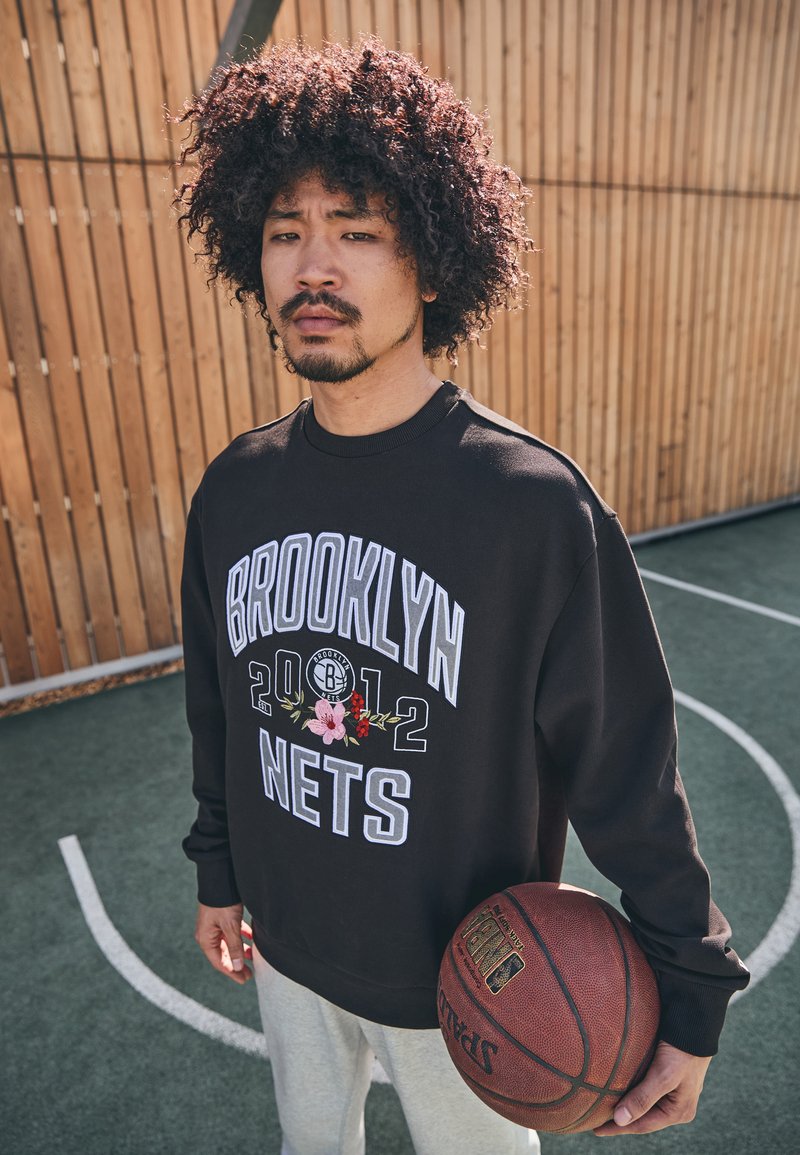 Felpa nera con "BROOKLYN NETS 2012" in bianco e grafiche floreali. Tiene un pallone da basket, indossa pantaloni grigi chiari, su uno sfondo di campo da basket.