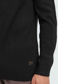 Black knit sweater featuring ribbed cuffs and hem, with a small logo patch on the lower left side. Soft texture, classic fit.