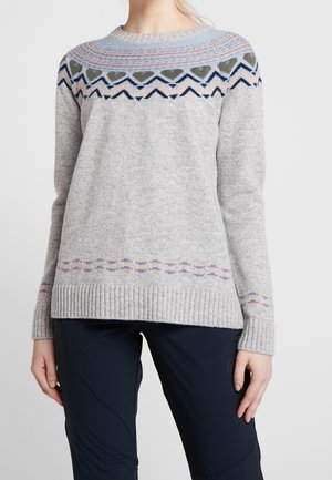 Vrouw draagt een lichtgrijze gebreide trui met blauwe, roze en olijfgroene hartjes en zigzagpatronen aan de pasvorm en zoom, gecombineerd met een donkere broek.