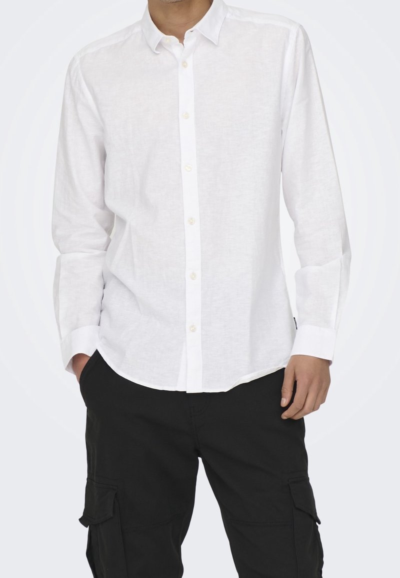 Homme portant une chemise blanche à manches longues boutonnée et un pantalon cargo noir, debout les mains dans les poches devant un fond uni.