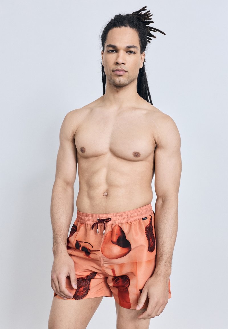 Homme torse nu avec de longues dreadlocks portant un short de bain orange avec des motifs abstraits noirs et rouges, debout devant un fond clair uni.