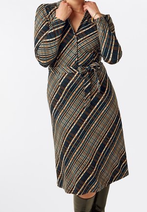 Femme portant une robe à manches longues à carreaux avec une ceinture à la taille, tenant le col près du cou, et portant un bracelet et une bague en or.