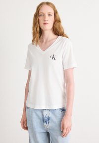 Balta medvilninė marškinėliai su V formos iškirpte ir trumpomis rankovėmis, kairėje krūtinės pusėje yra mažas juodas Calvin Klein logotipas. Laisvas kirpimas.