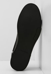 Semelle de chaussure noire avec surface en caoutchouc texturée. Présente un design de bord lisse et un logo "Calvin Klein" estampillé. Fermeture éclair latérale visible.