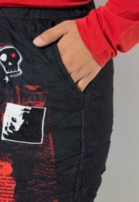 Schwarzer Stoffrock mit zerknitterter Textur, der rote Grafikdrucke zeigt, darunter ein Totenschädel und kontrastierende Bilder. Eine seitliche Tasche ist sichtbar.
