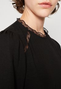 Mustapaita, jossa on pitsireunallinen kaula-aukko ja hillityt olkapleatit. Kangas on kevyt ja sileätekoinen.