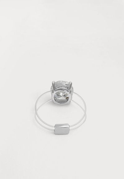 Anillo de plata con una gran piedra preciosa redonda y transparente engastada en garras, que cuenta con una banda doble delgada conectada por un cierre rectangular.