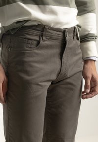 Pantalones de algodón en un tono verde oliva apagado, que cuentan con un bolsillo frontal, cierre de botón y una textura suave. Diseño liso con un detalle mínimo.