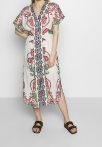 Biała lniana sukienka z kolorowym haftem paisley i kwiatowym, z dekoltem w kształcie litery V i krótkimi rękawami, sięgająca do połowy łydki.