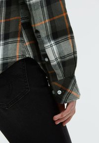 Grau-schwarzes karriertes Flanellhemd mit orangefarbenen Akzenten, mit geknöpften Manschetten, über schwarzen Denim-Jeans getragen. Nahaufnahme des Ärmels und der Hand.