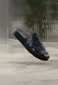 Μαύρο ανοιχτό σανδάλι slide με φαρδιές δερμάτινες λουριά και υφή στην σόλα, παρουσιασμένο μπροστά σε θολό σύγχρονο εσωτερικό φόντο.