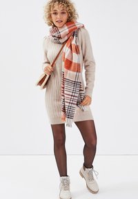 Vestido de punto beige con detalle de trenzado, bufanda de cuadros naranja y marrón, medias negras y botines crema con diseño de cordones.