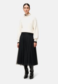 Weißer, flauschiger Pullover mit hohem, gerafftem Kragen, kombiniert mit einem schwarzen, plissierten Tüllrock. Lange schwarze Lederstiefel mit spitzem Schuhabschluss runden das Outfit ab.