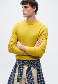 Jeune homme les bras croisés portant un pull texturé jaune et une chemise à carreaux bleus nouée autour de la taille, regardant vers la gauche sur un fond uni.